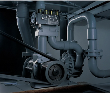 Close-up view of an industrial elevator hydraulic pump system with pipes, valves, and a drive belt mechanism in a dimly lit mechanical room.