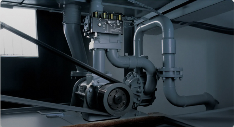 Close-up view of an industrial elevator hydraulic pump system with pipes, valves, and a drive belt mechanism in a dimly lit mechanical room.
