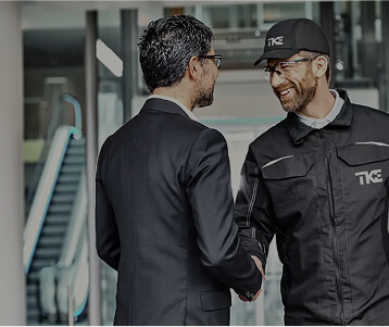 TKE technician in uniform shaking hands with a businessman in a modern building lobby, symbolizing partnership and service.