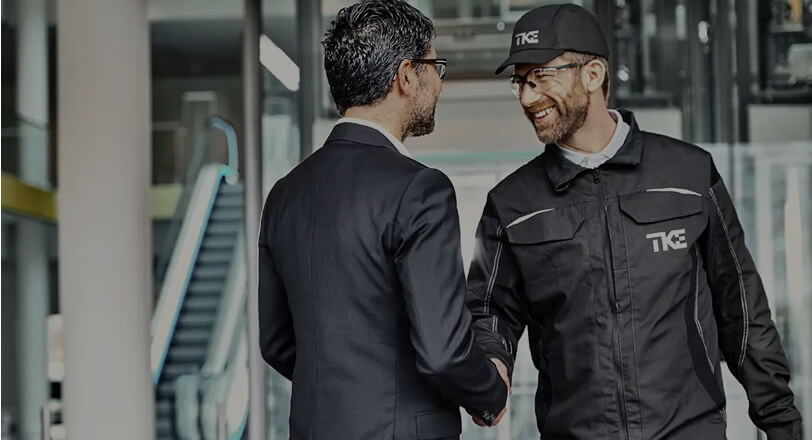 TKE technician in uniform shaking hands with a businessman in a modern building lobby, symbolizing partnership and service.